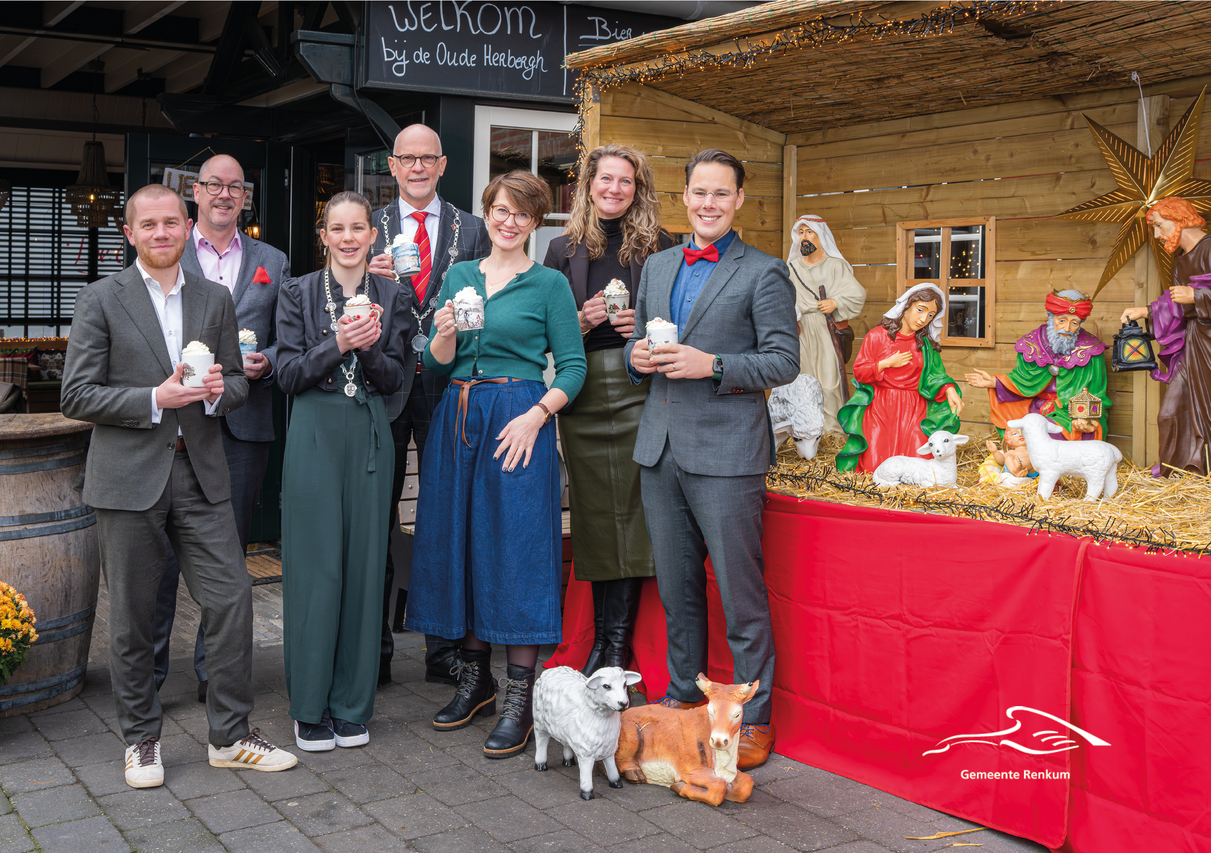 College van burgemeester en wethouders bij een kerststal met een kop warme chocolademelk in de hand
